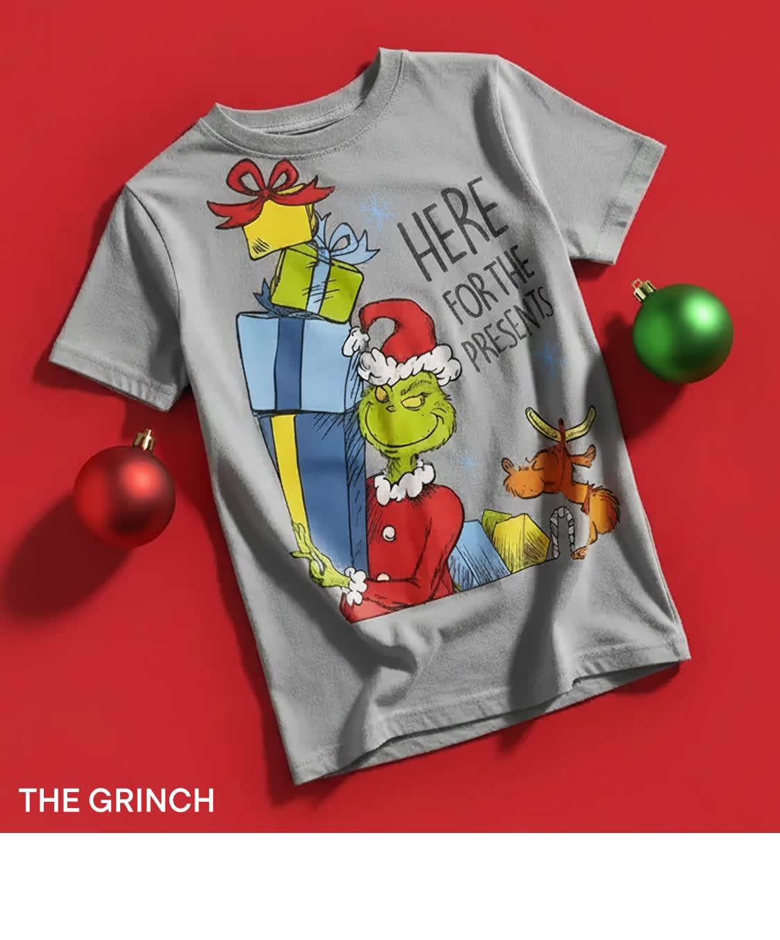 Un t-shirt gris orné d'une illustration colorée du personnage du Grinch entouré de cadeaux et de décorations de Noël sur un fond rouge vif.