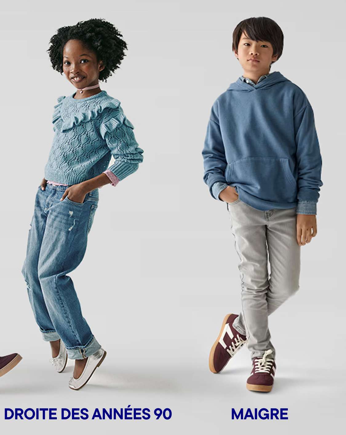 Two children, a girl with curly hair and a boy, are standing in a plain white background. The girl is wearing a light blue sweater and jeans, while the boy is wearing a blue hoodie and light-colored pants.