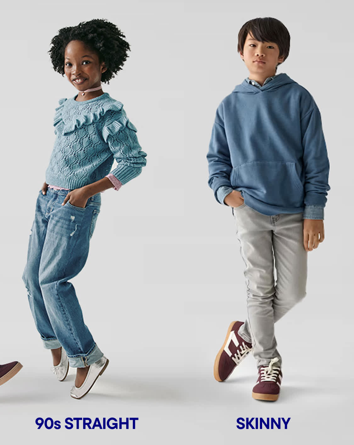 Two children, a girl with curly hair and a boy, are standing in a plain white background. The girl is wearing a light blue sweater and jeans, while the boy is wearing a blue hoodie and light-colored pants.