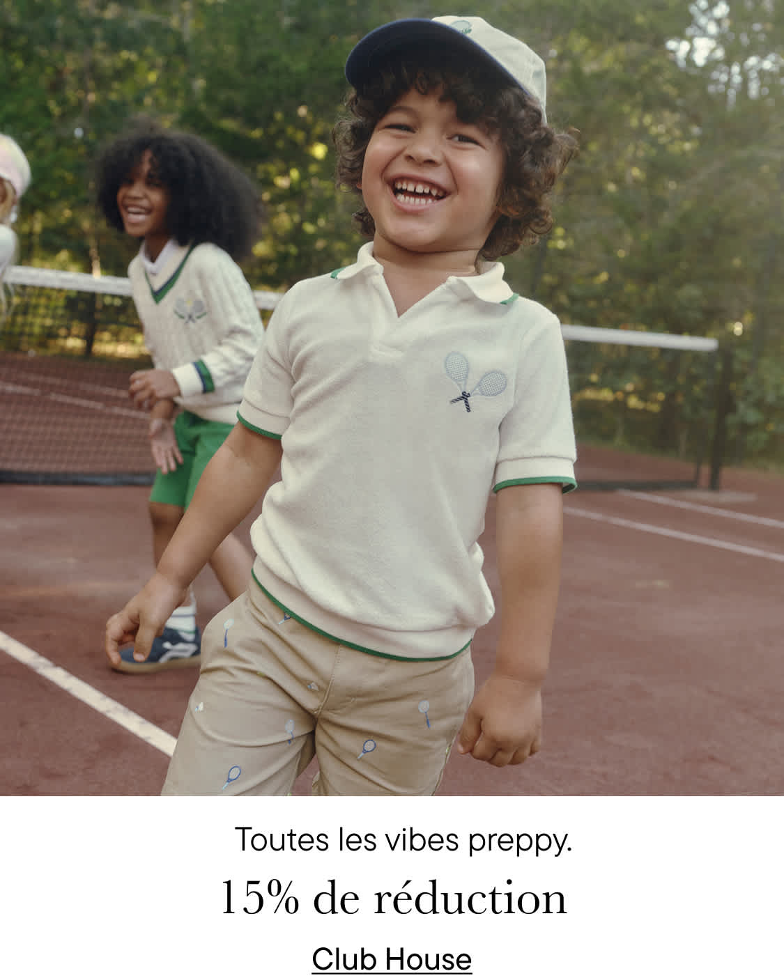 Deux jeunes enfants, un garçon et une fille, sourient et jouent sur un court de tennis entouré d'une pelouse parsemée de fleurs jaunes.