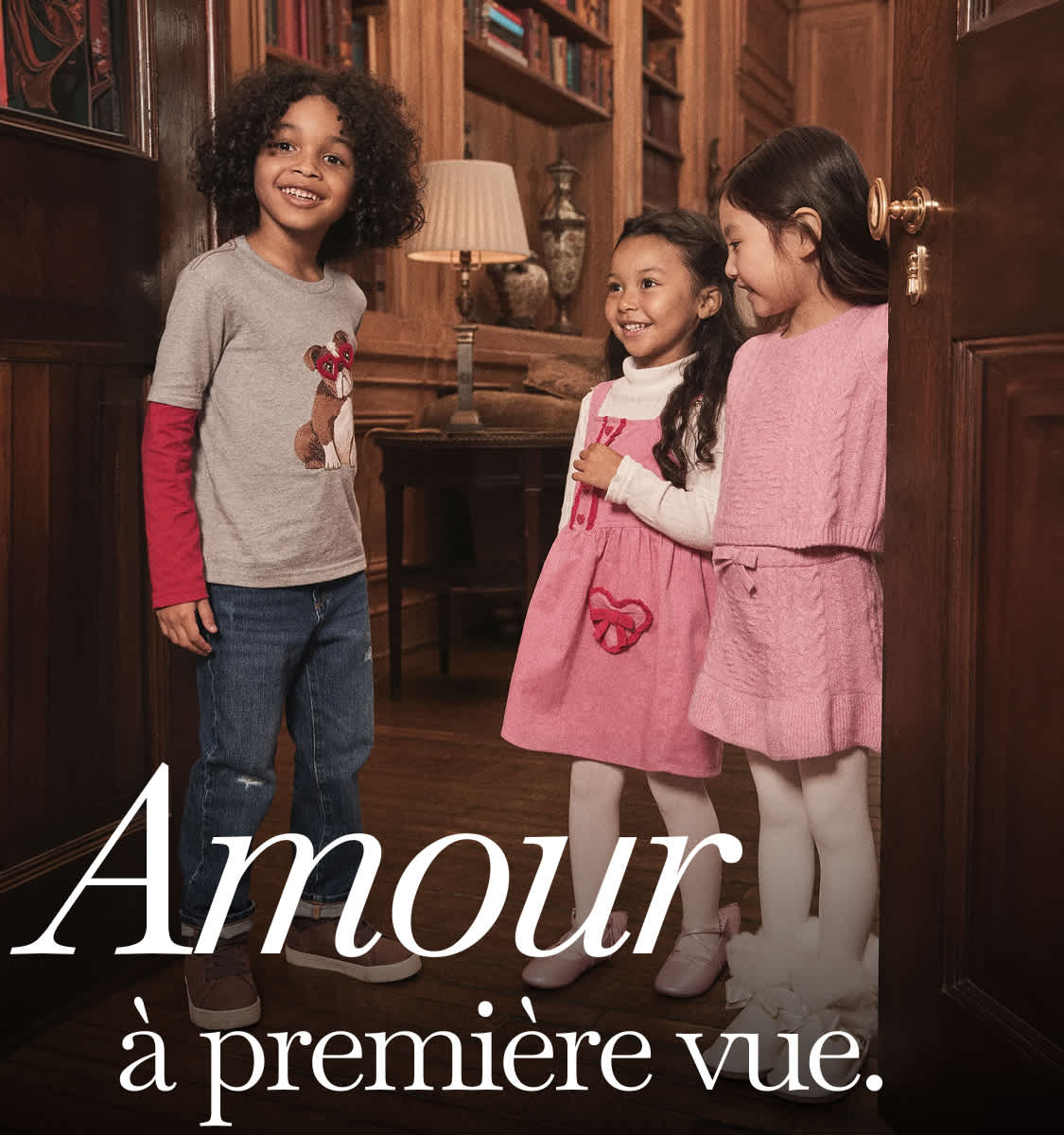 Trois jeunes filles, l'une aux cheveux bouclés et les autres aux cheveux lisses, se tiennent ensemble dans une pièce confortable aux murs lambrissés, avec des étagères à livres en arrière-plan, créant une atmosphère chaleureuse et intime.