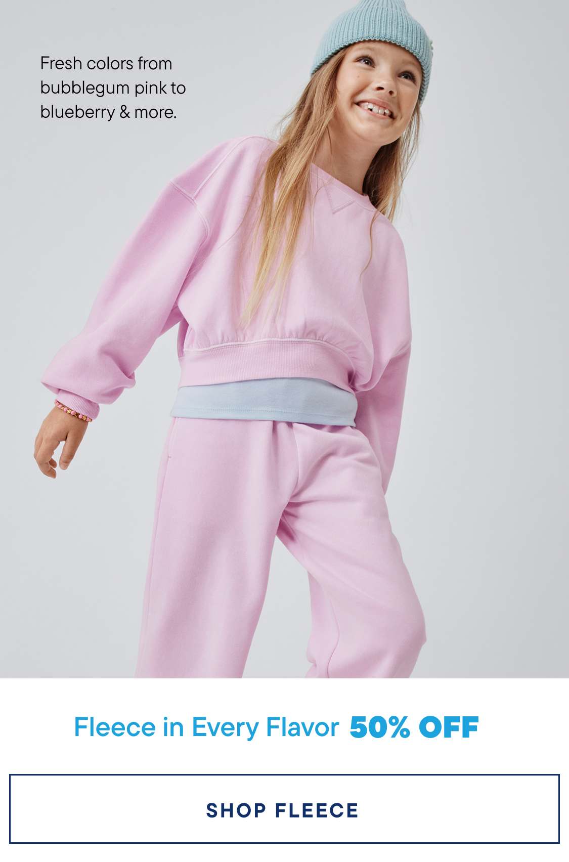 A young woman with long blonde hair is wearing a pink sweatsuit and a light blue beanie, smiling at the camera against a plain white background.