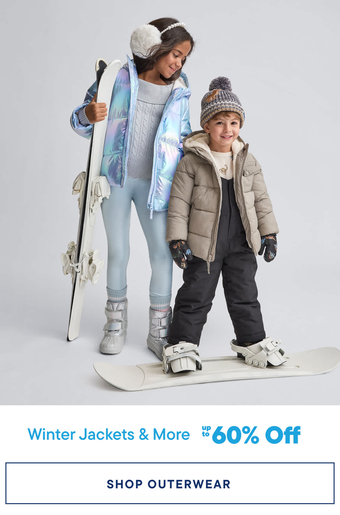 Two young children, a girl and a boy, are dressed in warm winter clothing and holding ski equipment, standing against a plain white background.