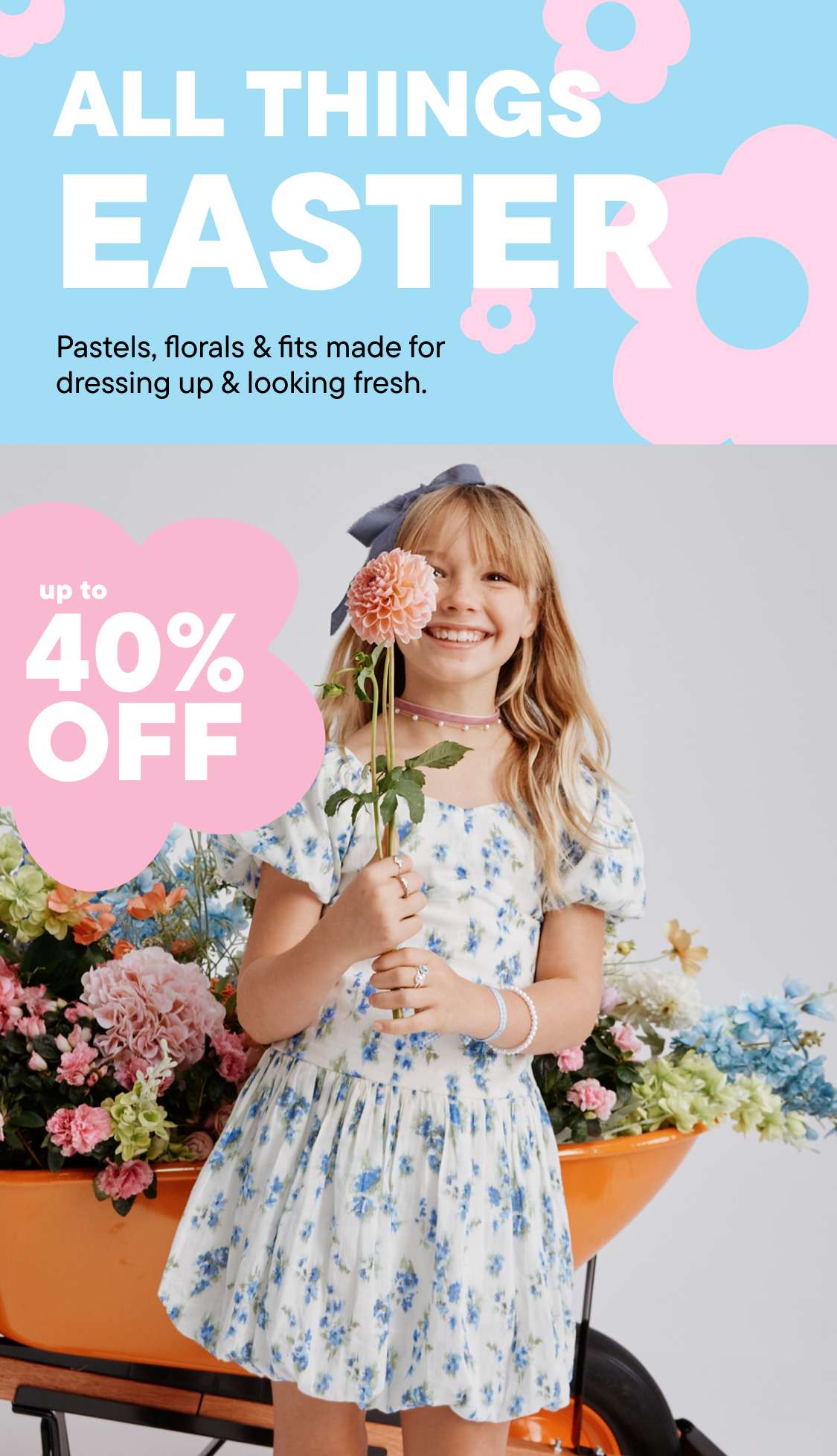 A young woman with curly blonde hair is smiling and holding a bouquet of flowers in front of a colorful floral background.