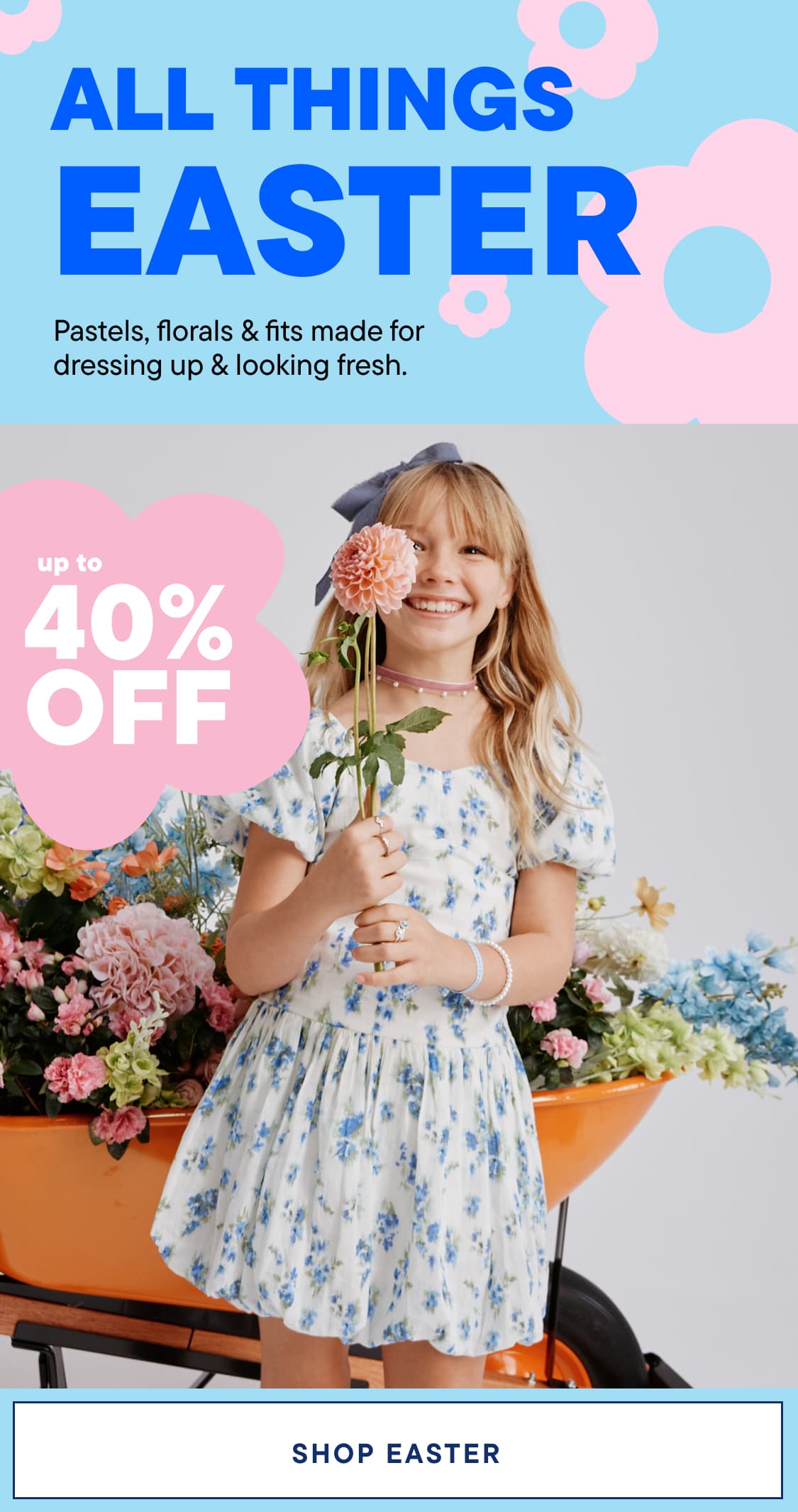 A young woman with curly blonde hair is smiling and holding a bouquet of flowers in front of a colorful floral background.