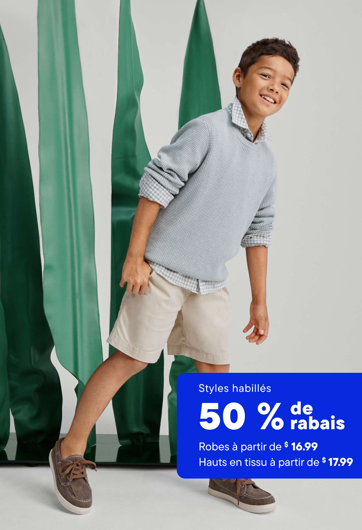 Un jeune garçon vêtu d'un pull gris et d'un short beige se tient devant un fond vert vif, sur lequel est affichée une banderole publicitaire annonçant une réduction de 50 % sur les vêtements habillés.