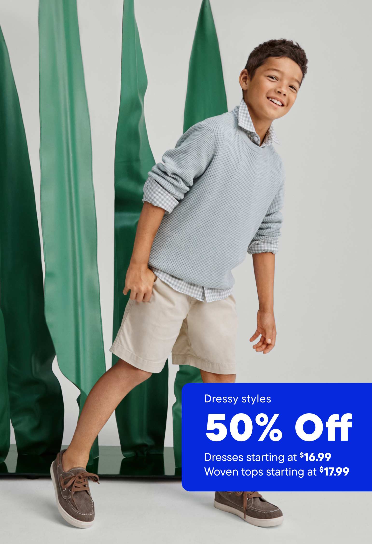 A young boy wearing a gray sweater and beige shorts stands in front of a vibrant green backdrop, with a promotional banner advertising a 50% off sale on dressy styles.