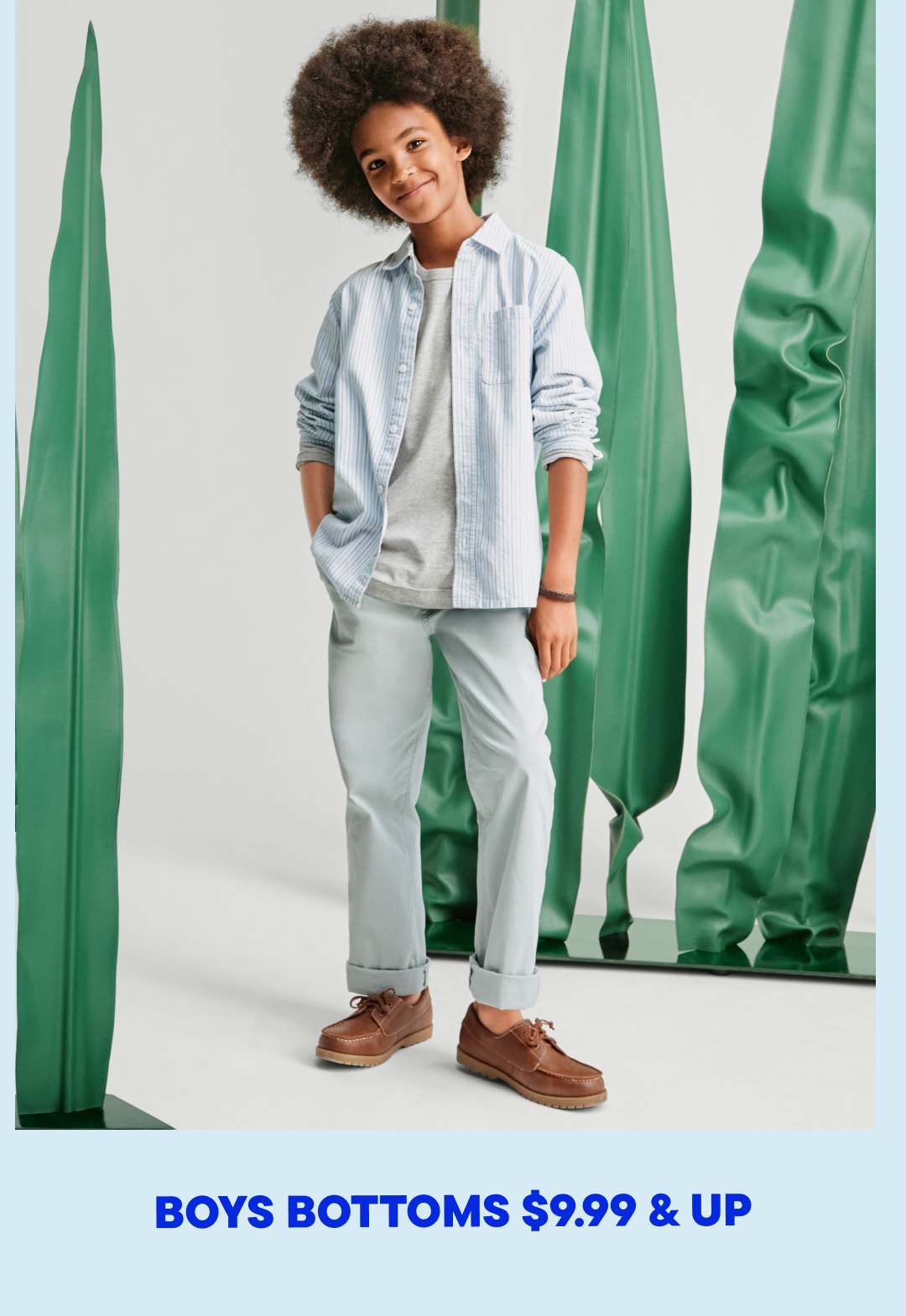 A young person with curly hair wearing a light blue jacket, gray pants, and brown shoes stands in front of a green backdrop.