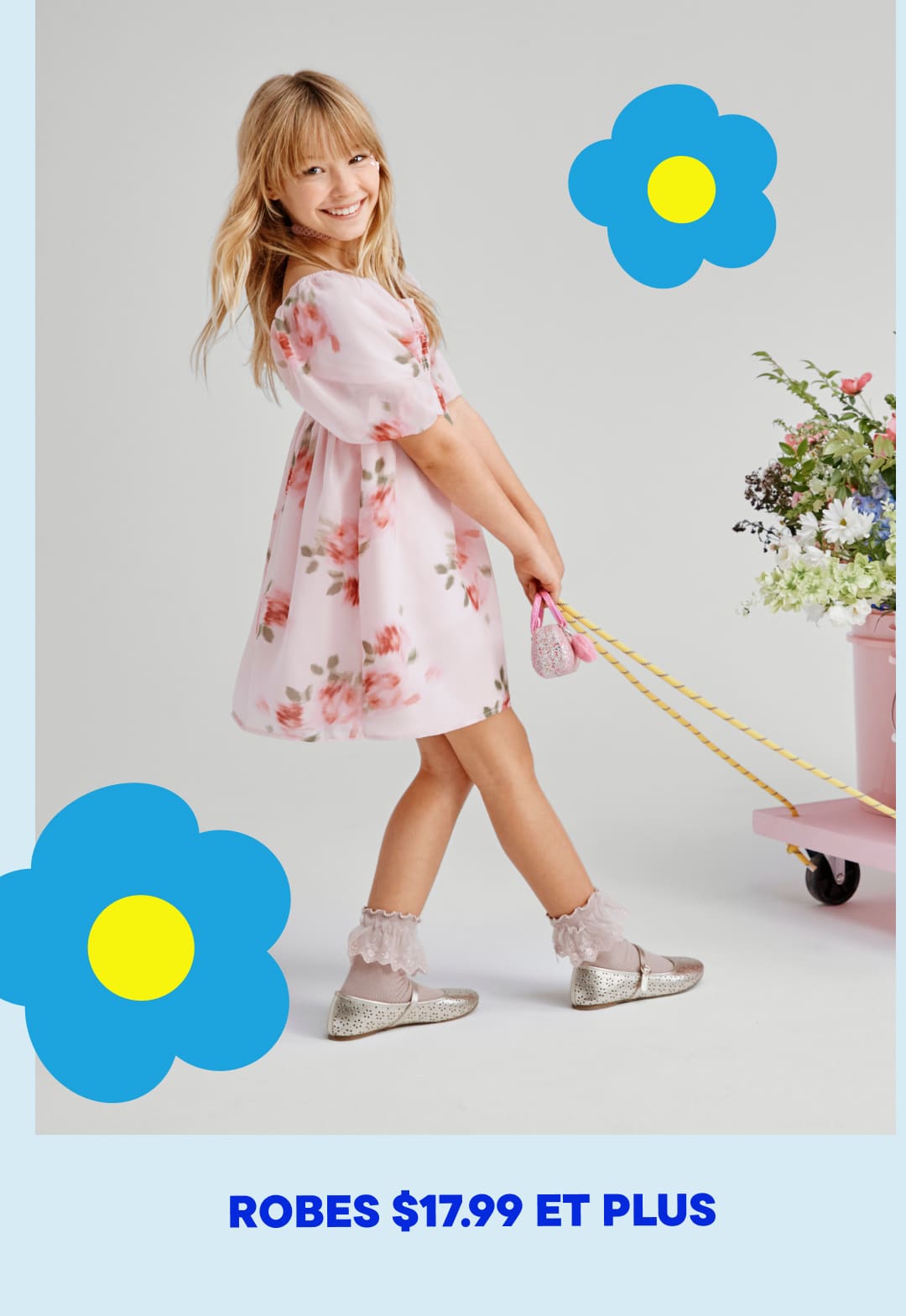 Une jeune fille vêtue d'une robe rose à fleurs se tient debout, souriante, avec des fleurs bleues et jaunes en arrière-plan.