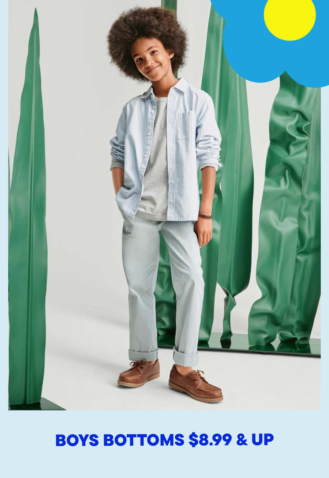 A young person with curly hair wearing a light blue jacket, gray pants, and brown shoes stands in front of a plain white background with large green fabric panels on either side.