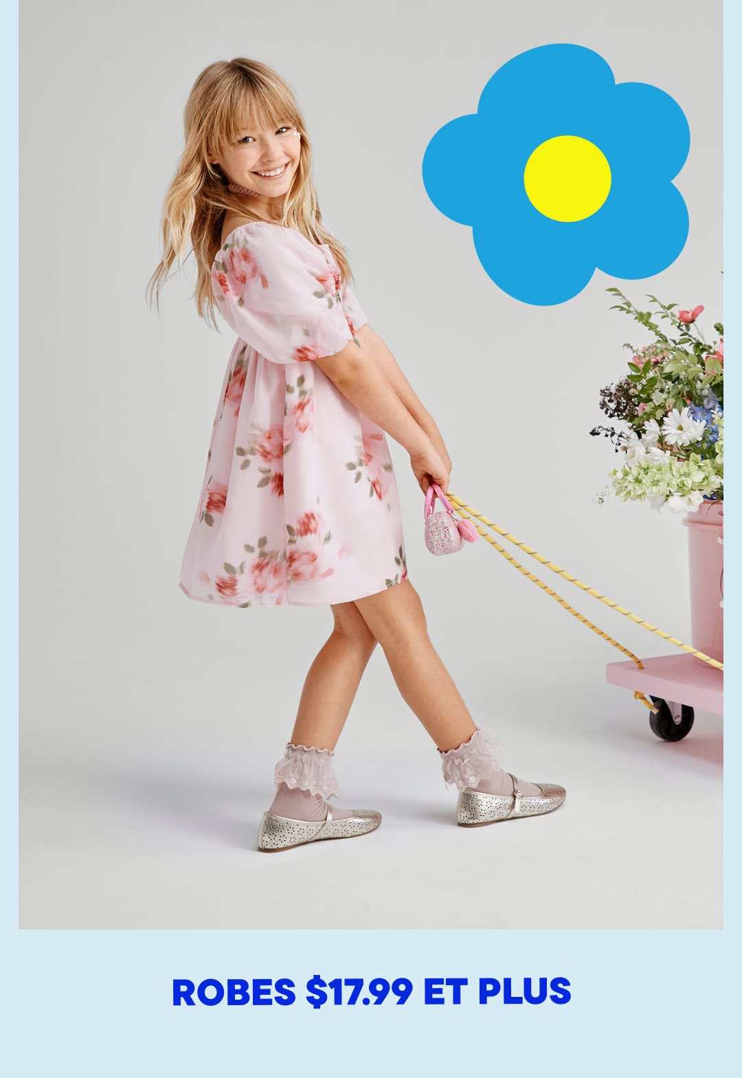 Une jeune fille vêtue d'une robe rose à fleurs se tient devant un fond blanc, tenant un gros ballon bleu et jaune en forme de fleur.