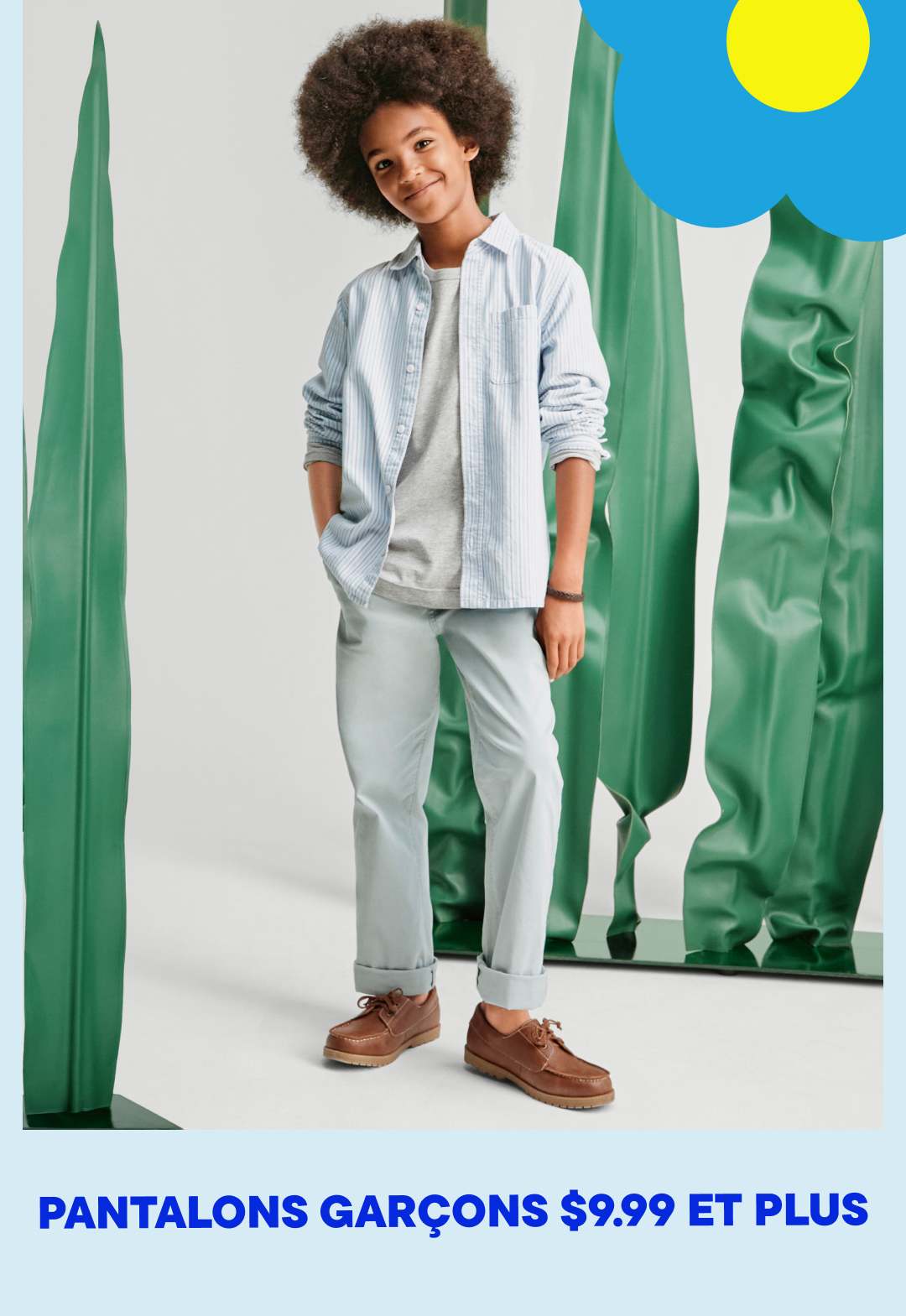 Une jeune personne aux cheveux bouclés, vêtue d'une veste bleu clair, d'un pantalon gris et de chaussures marron, se tient devant un fond blanc uni, encadré de grands panneaux de tissu vert.