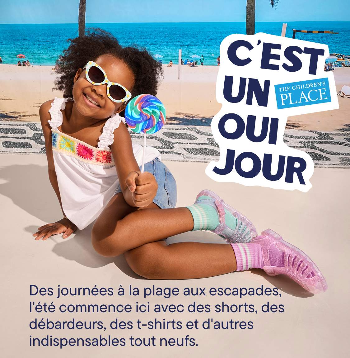 L'image montre deux jeunes enfants, une fille et un garçon, debout devant un panneau indiquant « C'EST LA JOURNÉE DU OUI » sur une plage ensoleillée avec des palmiers en arrière-plan.