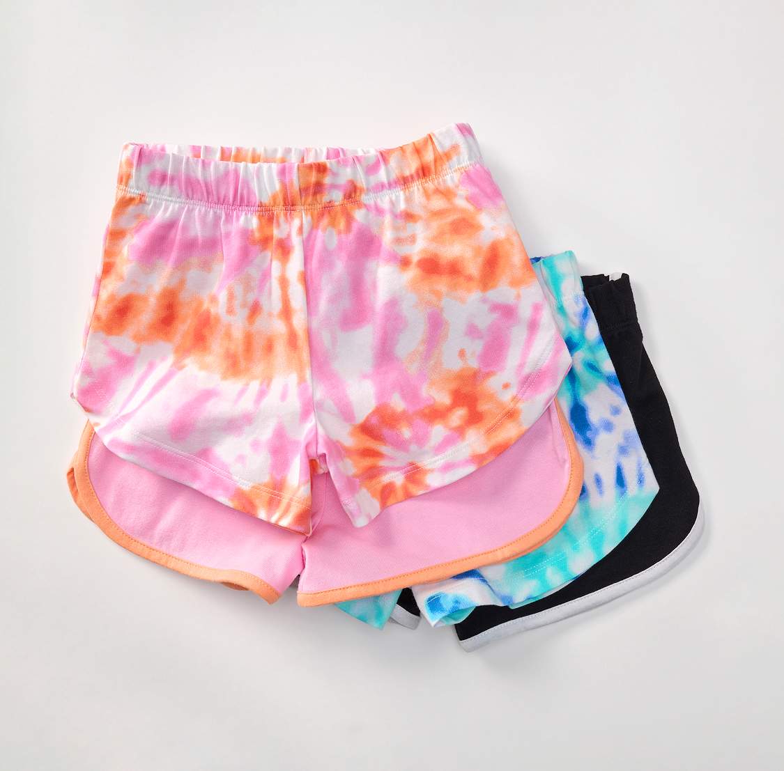 Three pairs of colorful shorts, with a tie-dye pair in the foreground and two solid colored pairs in the background, are laid out on a white surface.