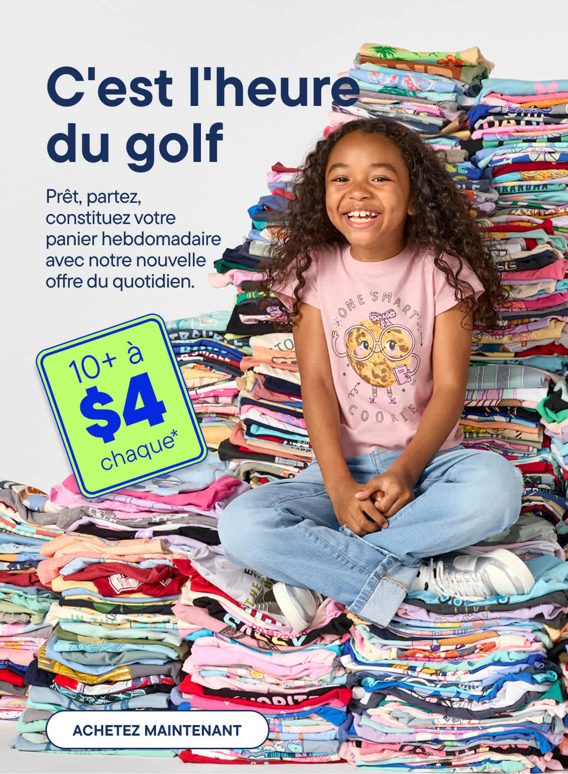 Une jeune fille souriante est assise en tailleur devant une énorme pile de t-shirts pliés et colorés, avec une pancarte verte annonçant une promotion au premier plan.