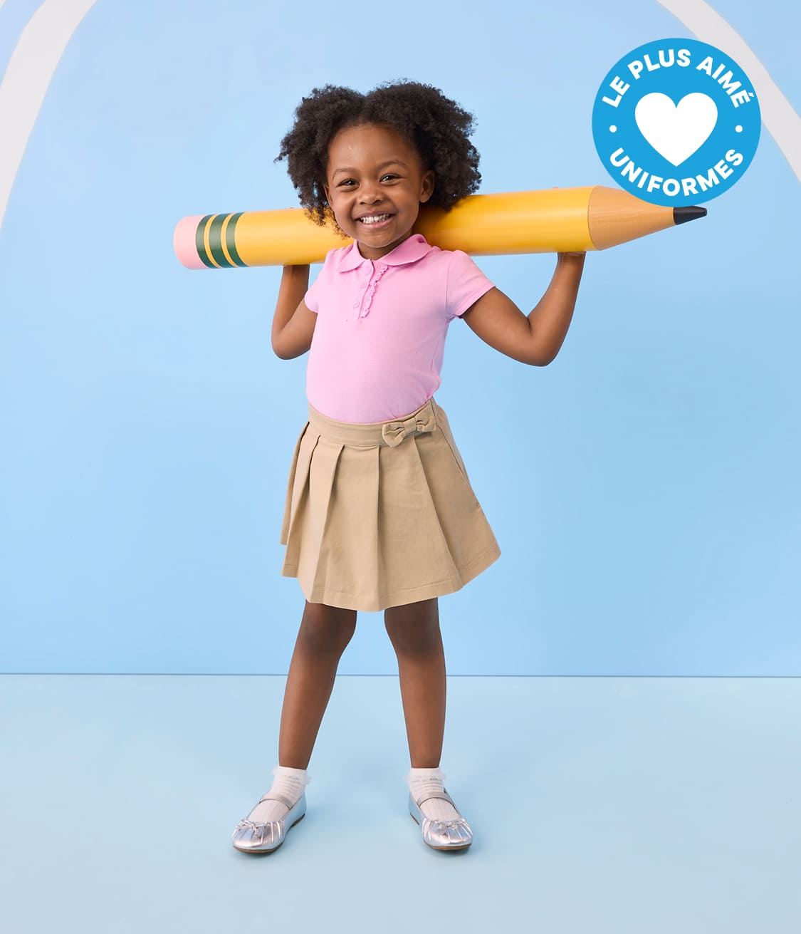 Une jeune fille souriante, vêtue d'un polo rose et d'une jupe kaki, tient un crayon jaune géant sur ses épaules, sur un fond bleu clair à rayures blanches.