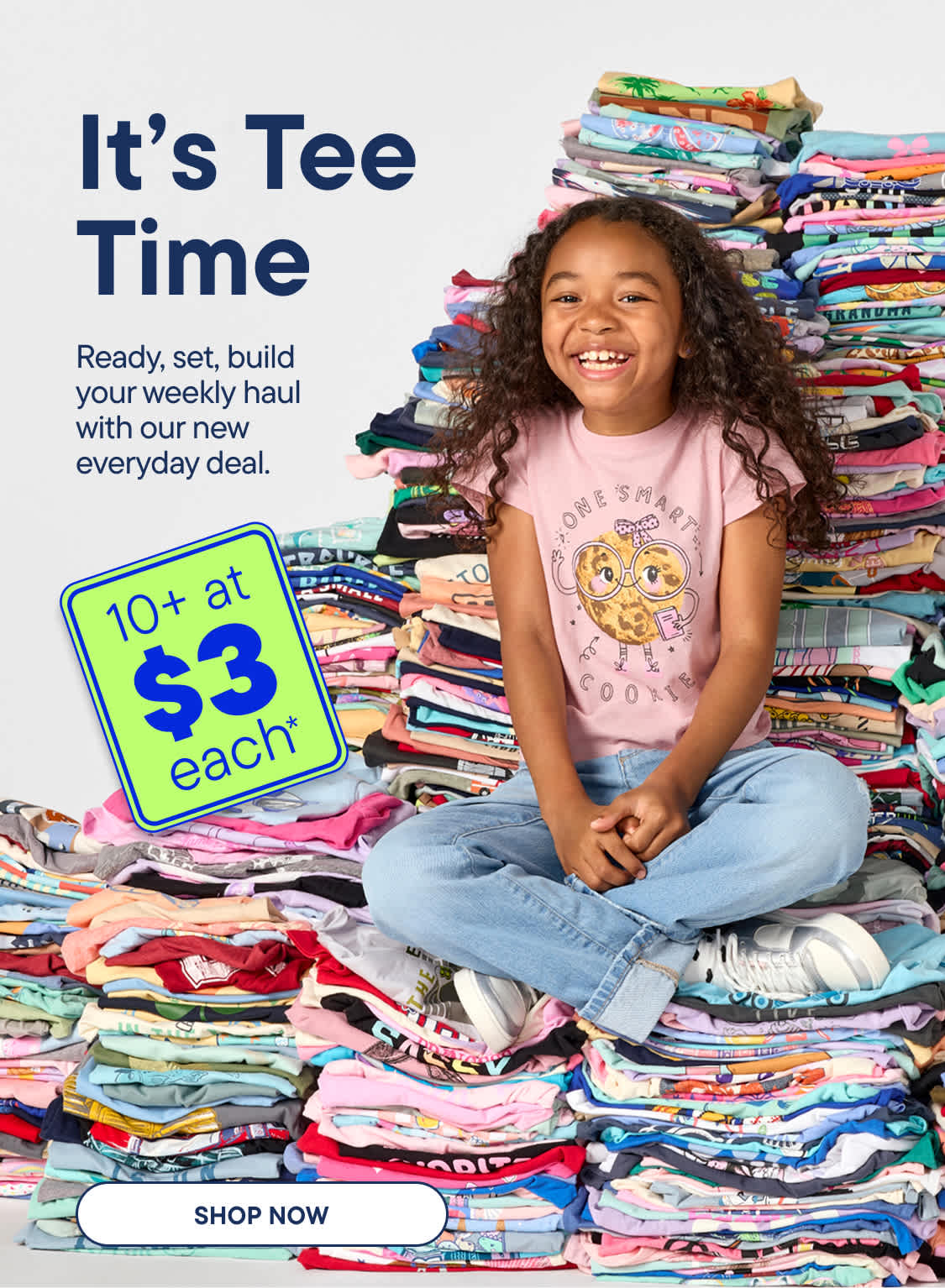 A smiling young girl sits cross-legged in front of a massive, colorful pile of folded t-shirts, with a sign advertising 10+ at $3 each to her left.