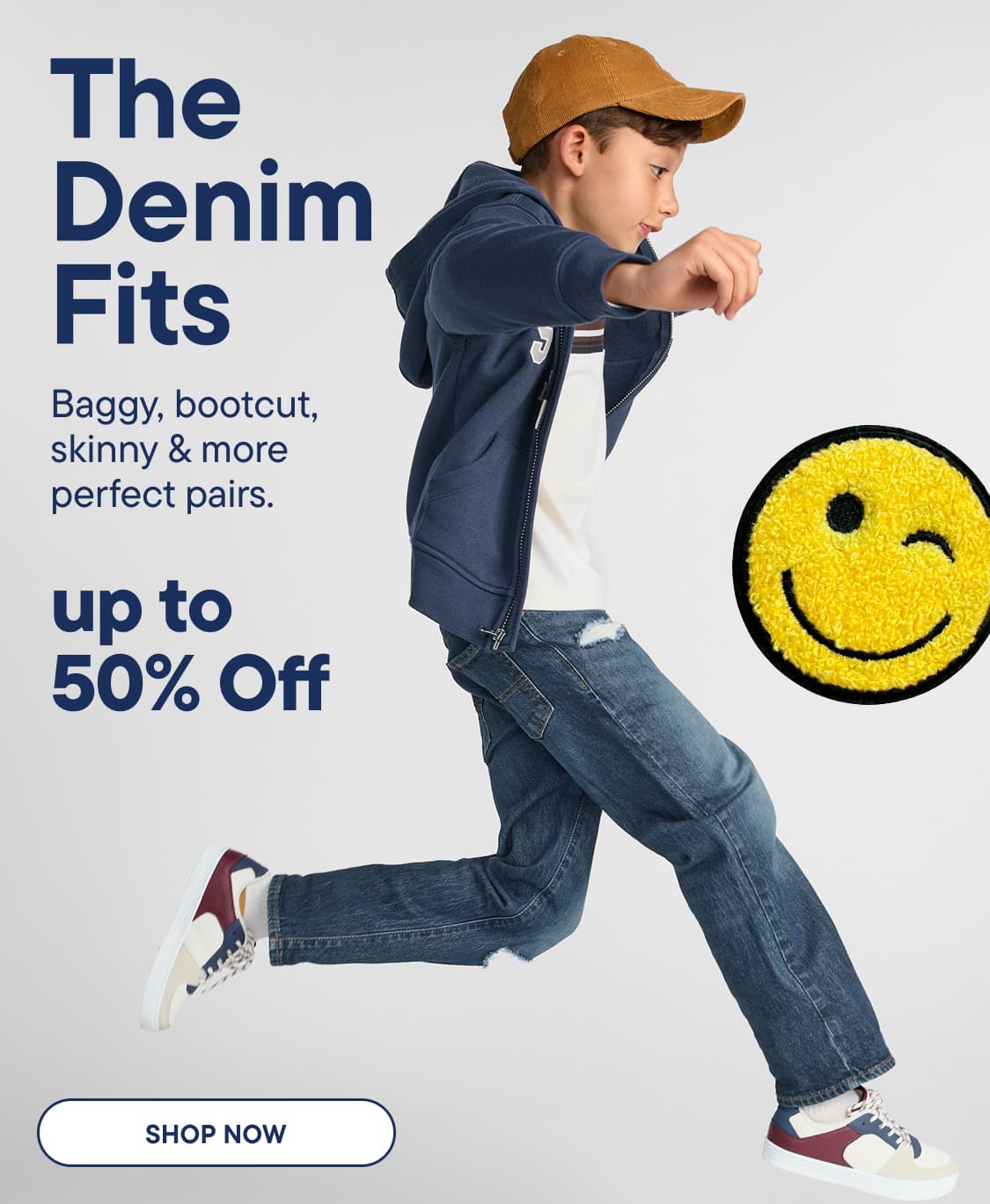 A young boy in denim jeans and a hoodie jumps in the foreground, with a winking smiley face and sale text on a light gray background | THE DENIM FITS UP TO 50% OFF