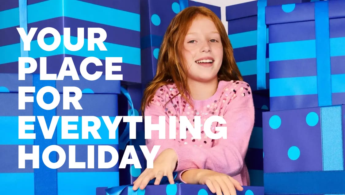 A smiling girl in a pink sweater sits in the foreground, surrounded by large blue and purple wrapped gift boxes in the background.