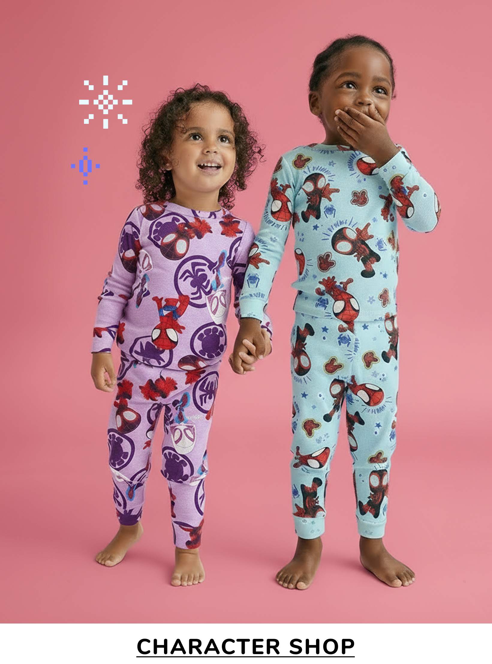 Two young children, a girl and a boy, are standing together in colorful pajamas against a pink background. The girl is wearing a purple outfit with a floral pattern, while the boy is wearing a blue outfit with a bear pattern. They are both smiling and appear to be having fun.