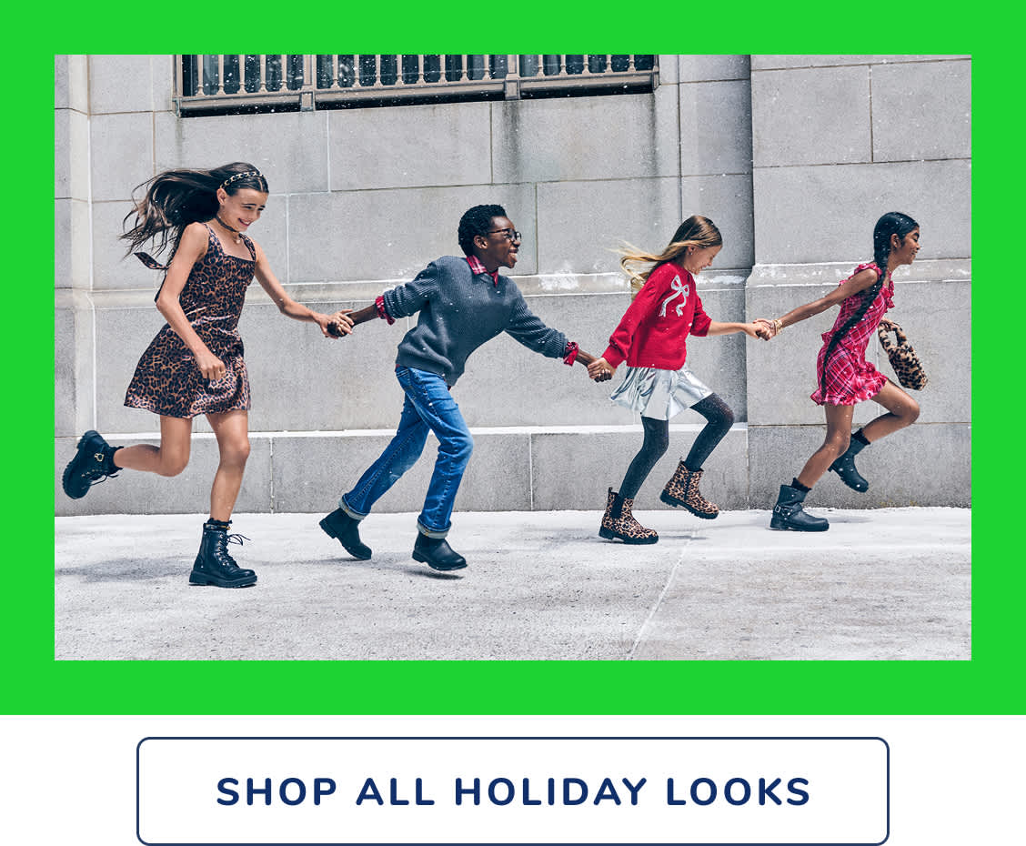 Four fashionably dressed individuals, wearing vibrant and eclectic outfits, are walking together in front of a plain gray concrete wall backdrop.