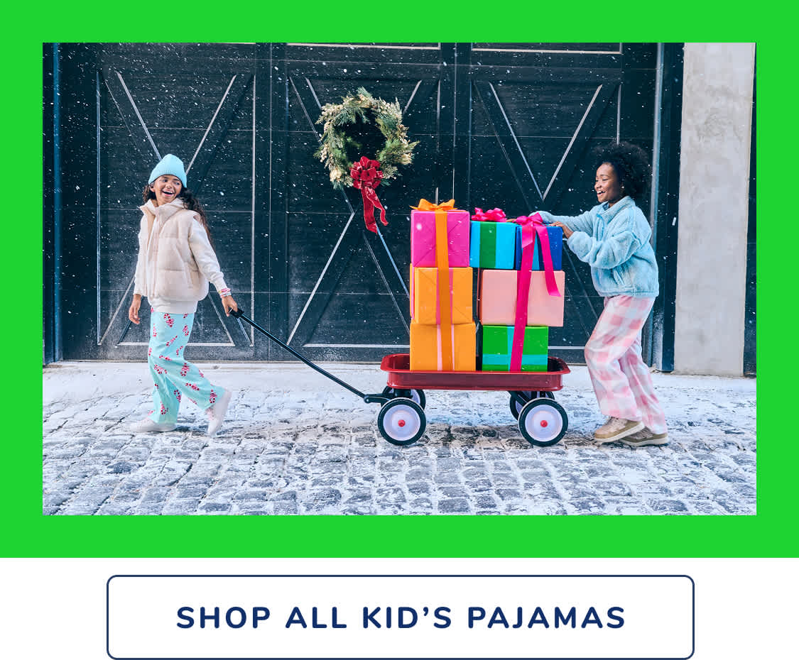 Two individuals in colorful winter attire stand in front of a storefront window, with a cart full of vibrant gift boxes and a festive wreath hanging above.