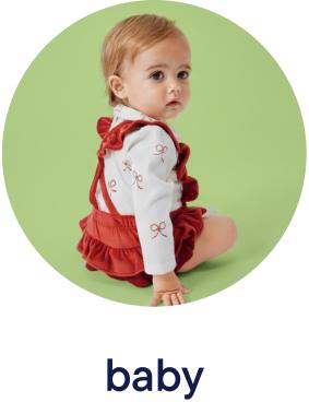 A young baby girl wearing a red and white ruffled dress sits on a green background, looking directly at the camera with a curious expression.