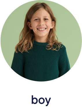 A smiling young girl with wavy brown hair wearing a green sweater against a light green background.