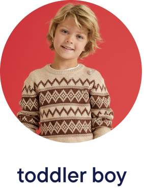 A young boy with blond, wavy hair is wearing a patterned sweater and smiling against a bright red background.