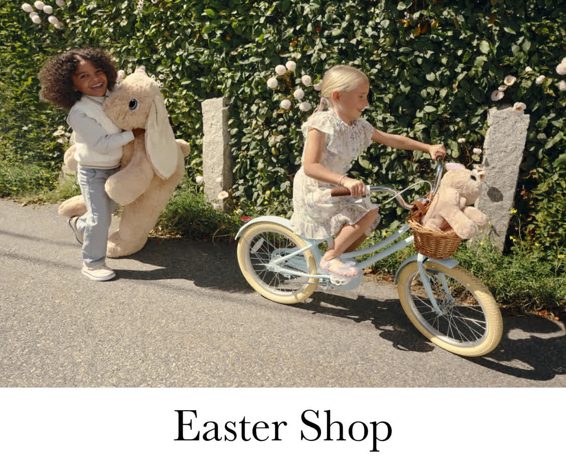 The image shows a young woman with curly hair wearing a light blue shirt, standing in a grassy field with a body of water in the background. Next to her is another young woman wearing a white sweater.