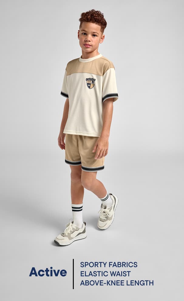 La imagen muestra a un niño pequeño con una camiseta y pantalones cortos de color beige, de pie sobre un fondo liso. Tiene una expresión amigable y parece formar parte de un anuncio de ropa deportiva de la marca "Active".