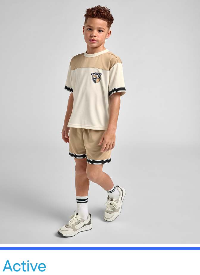 A young boy wearing a beige t-shirt and shorts, standing in a plain studio setting with a neutral background.