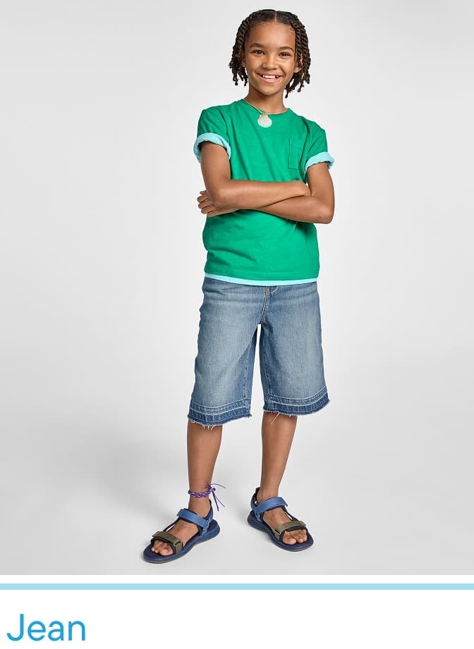 A young child with short, curly hair stands confidently in a green t-shirt, blue denim shorts, and sandals against a plain white background.