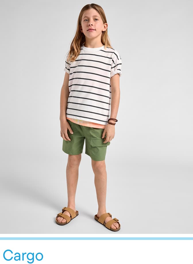 A young girl wearing a striped shirt and green shorts stands against a plain background, with sandals on her feet.