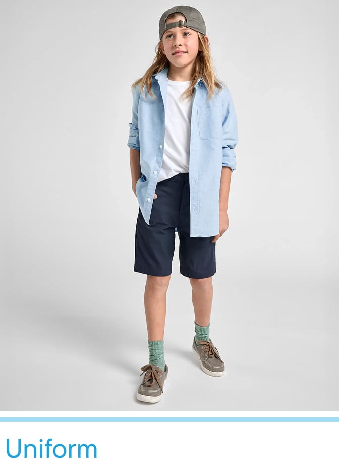 A young girl wearing a denim jacket, white shirt, and navy shorts stands against a plain white background, with her hands in her pockets and a cap on her head.