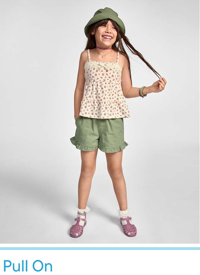 Una joven que viste una blusa con estampado floral, pantalones cortos verde oliva y sandalias aparece de pie sobre un fondo blanco liso, sonriendo y sosteniendo el ala de su sombrero verde.