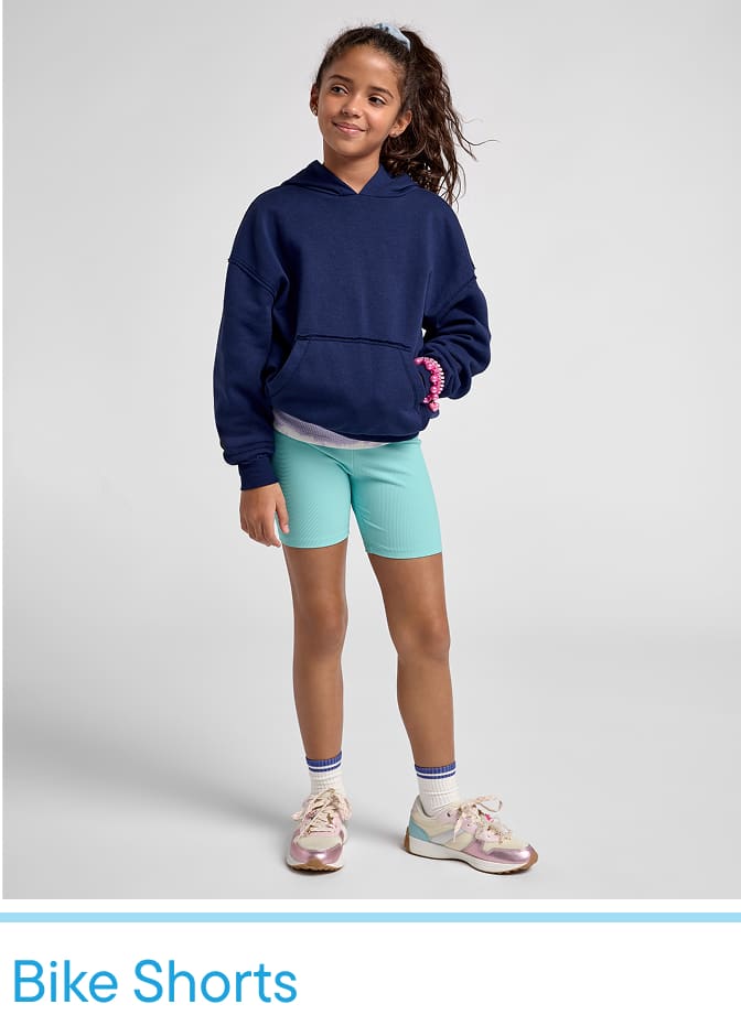 Una niña que viste una sudadera con capucha azul marino, pantalones cortos verde menta y zapatillas deportivas coloridas está parada frente a un fondo blanco liso.