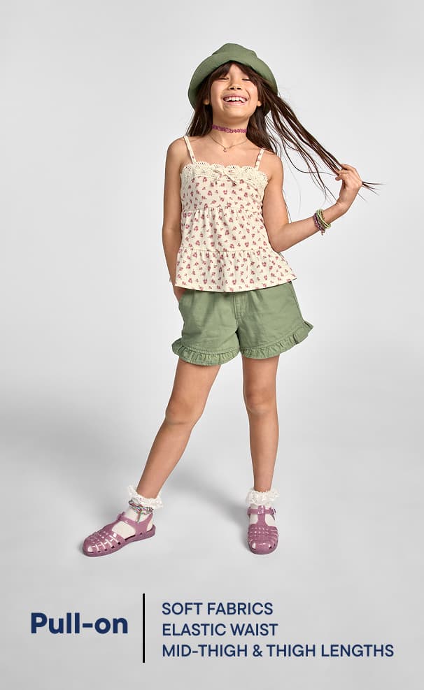 Una jovencita con una camiseta sin mangas estampada con flores, pantalones cortos verde oliva y sandalias rosas posa sobre un fondo blanco liso, con el pelo ondeando al viento y una radiante sonrisa en el rostro.