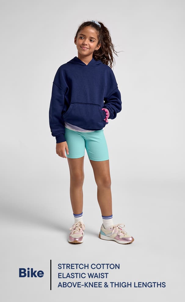 Una jovencita vestida con una sudadera azul marino, pantalones cortos verde menta y zapatillas deportivas de colores posa sobre un fondo blanco liso.