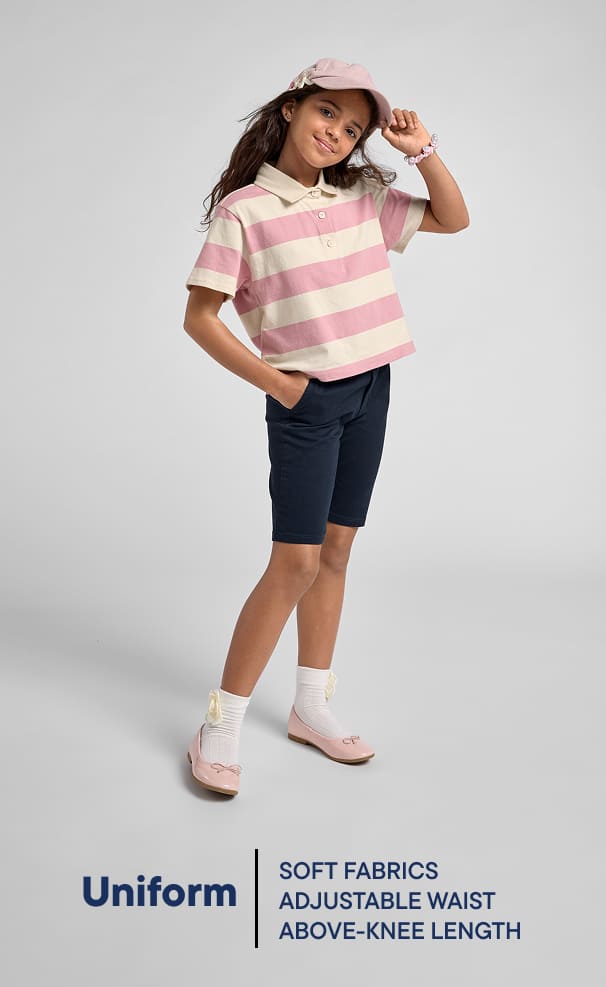 Una niña pequeña, vestida con una camiseta de rayas rosas y blancas, pantalones cortos azul marino y zapatos blancos, posa sobre un fondo blanco liso.