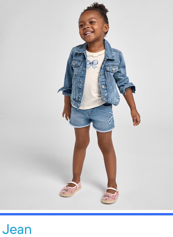 Un niño pequeño y sonriente, vestido con una chaqueta vaquera, una camisa blanca y pantalones cortos vaqueros, se encuentra frente a un fondo blanco liso.