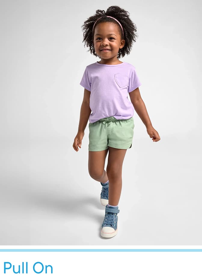 Un niño pequeño con cabello rizado que viste una camiseta lavanda, pantalones cortos verdes y zapatillas azules está parado sobre un fondo blanco liso.