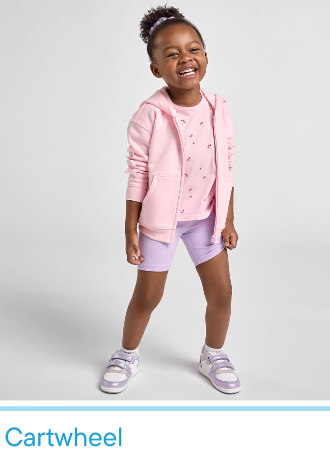 Una joven sonriente, vestida con una chaqueta rosa y pantalones cortos, aparece de pie frente a un fondo blanco liso.