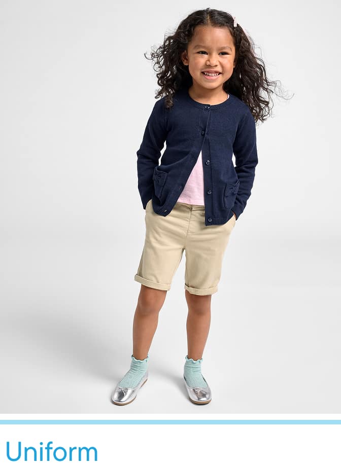 Una joven de cabello rizado, vestida con un cárdigan azul marino, pantalones cortos beige y zapatos plateados, aparece de pie frente a un fondo blanco liso, sonriendo alegremente.