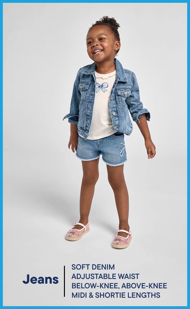A young, smiling child wearing a denim jacket, white shirt, and denim shorts stands against a plain white background.
