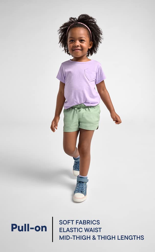 A young child with curly hair wearing a lavender shirt, green shorts, and blue sneakers stands against a plain white background.
