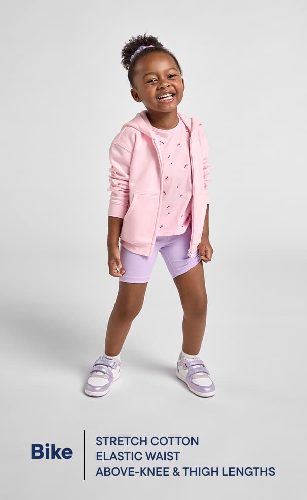 A young, smiling girl wearing a pink jacket and shorts stands against a plain white background.