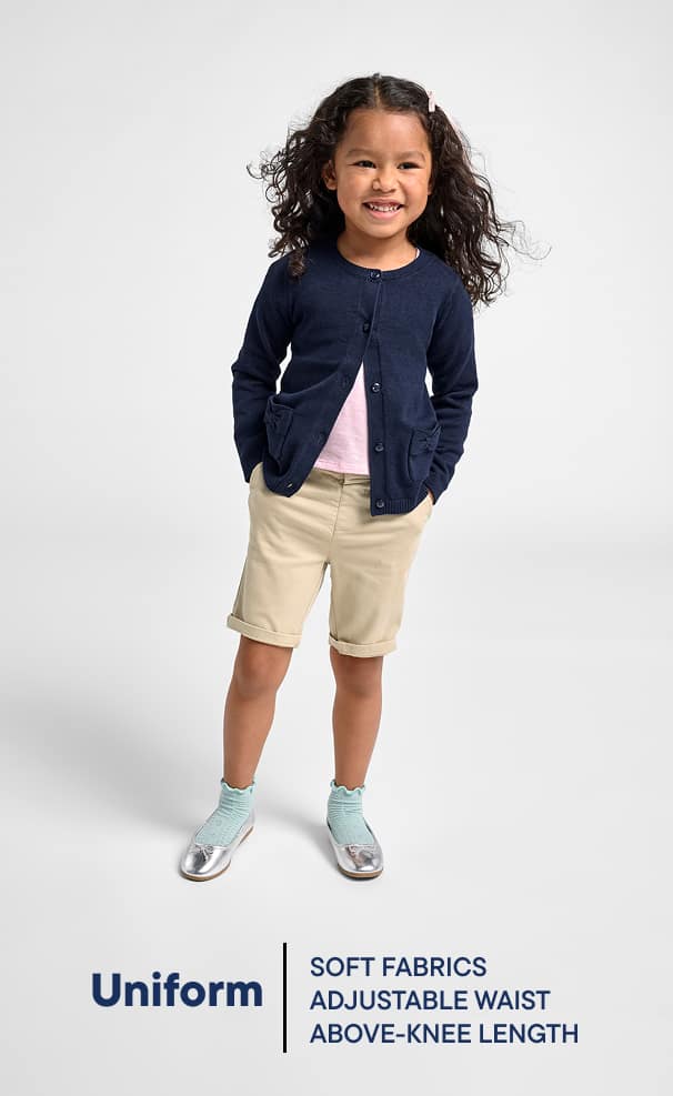 A young girl with curly hair wearing a navy blue cardigan, beige shorts, and silver shoes stands against a plain white background, smiling brightly.