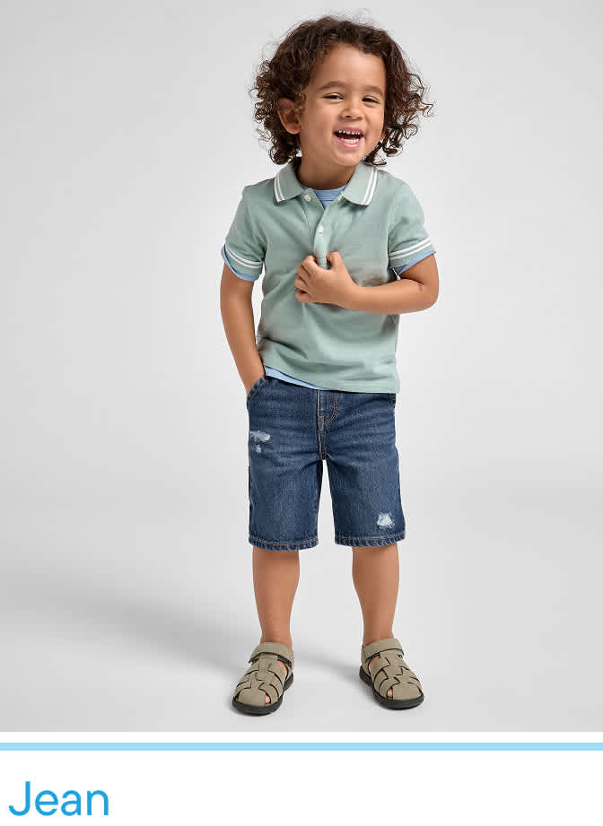 A young child with curly hair is standing and smiling, wearing a light green polo shirt and denim shorts, against a plain white background.