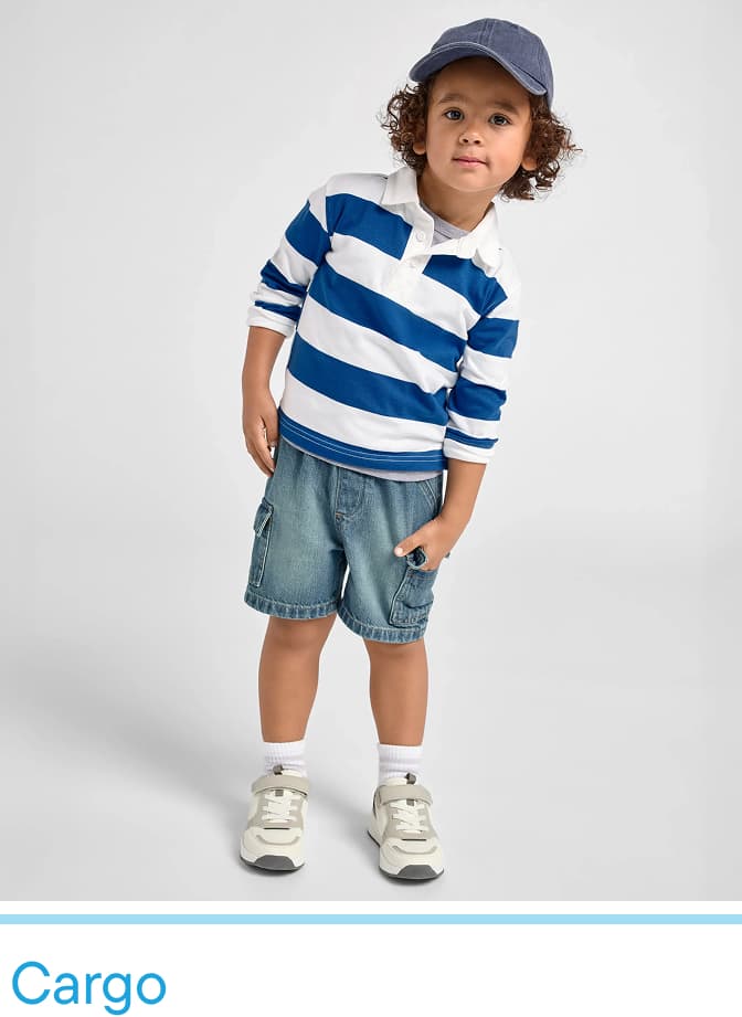 A young child wearing a striped shirt, denim shorts, and sneakers stands against a plain white background, with a cheerful expression on their face.